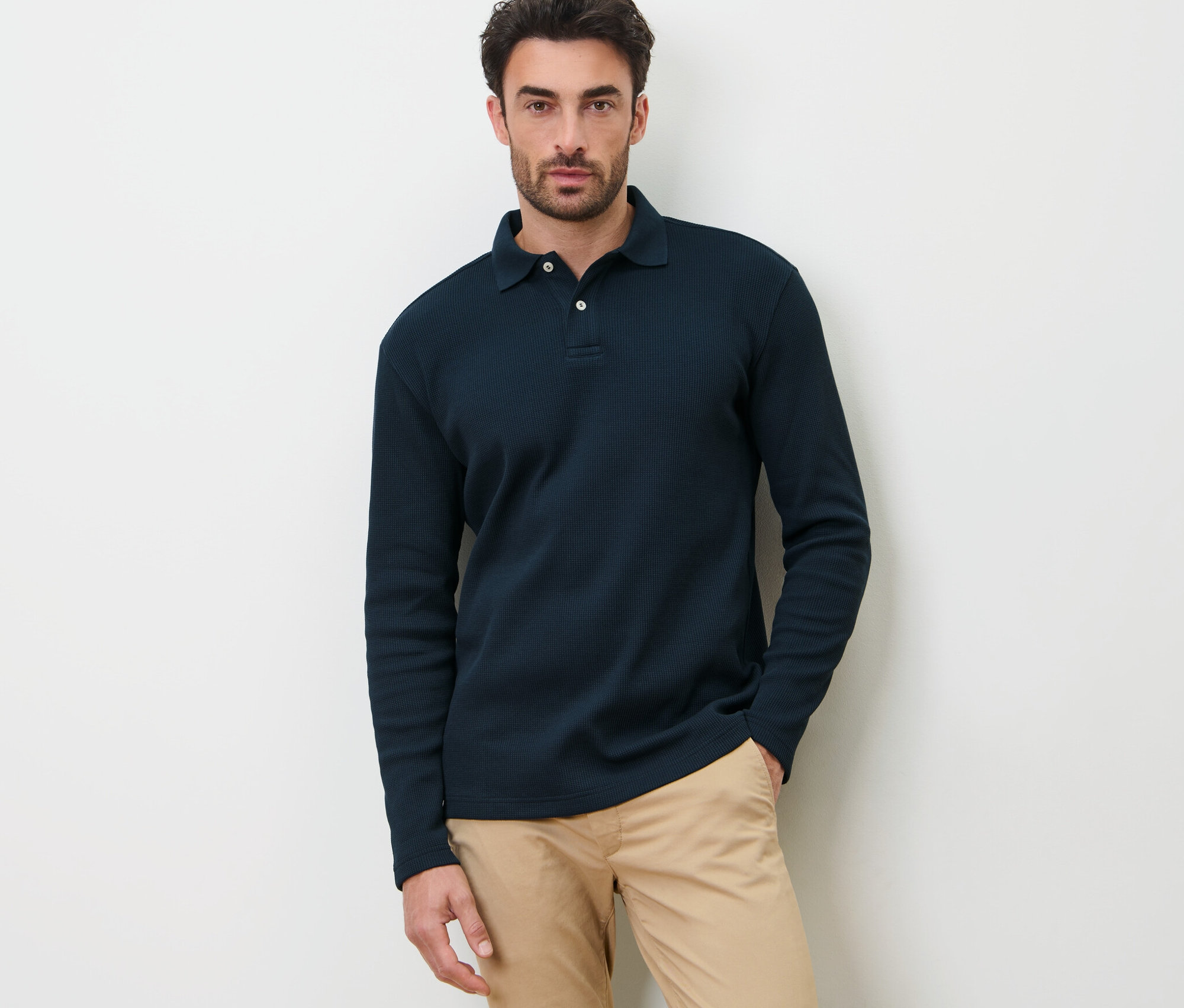 Egy férfi sötétkék, hosszú ujjú pólóingben pózol fehér háttér előtt.