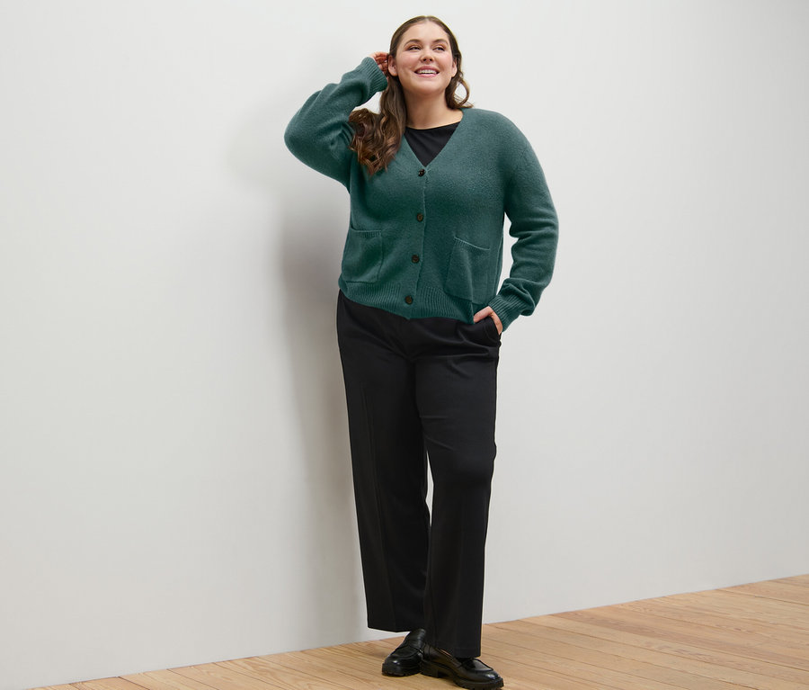 Nő pózol kötött, sötétzöld gyapjú kardigánban és fekete jersey nadrágban.