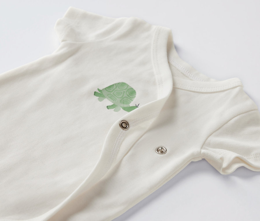 Egy fehér, rövid ujjú baba body közelképe, teknős mintával.