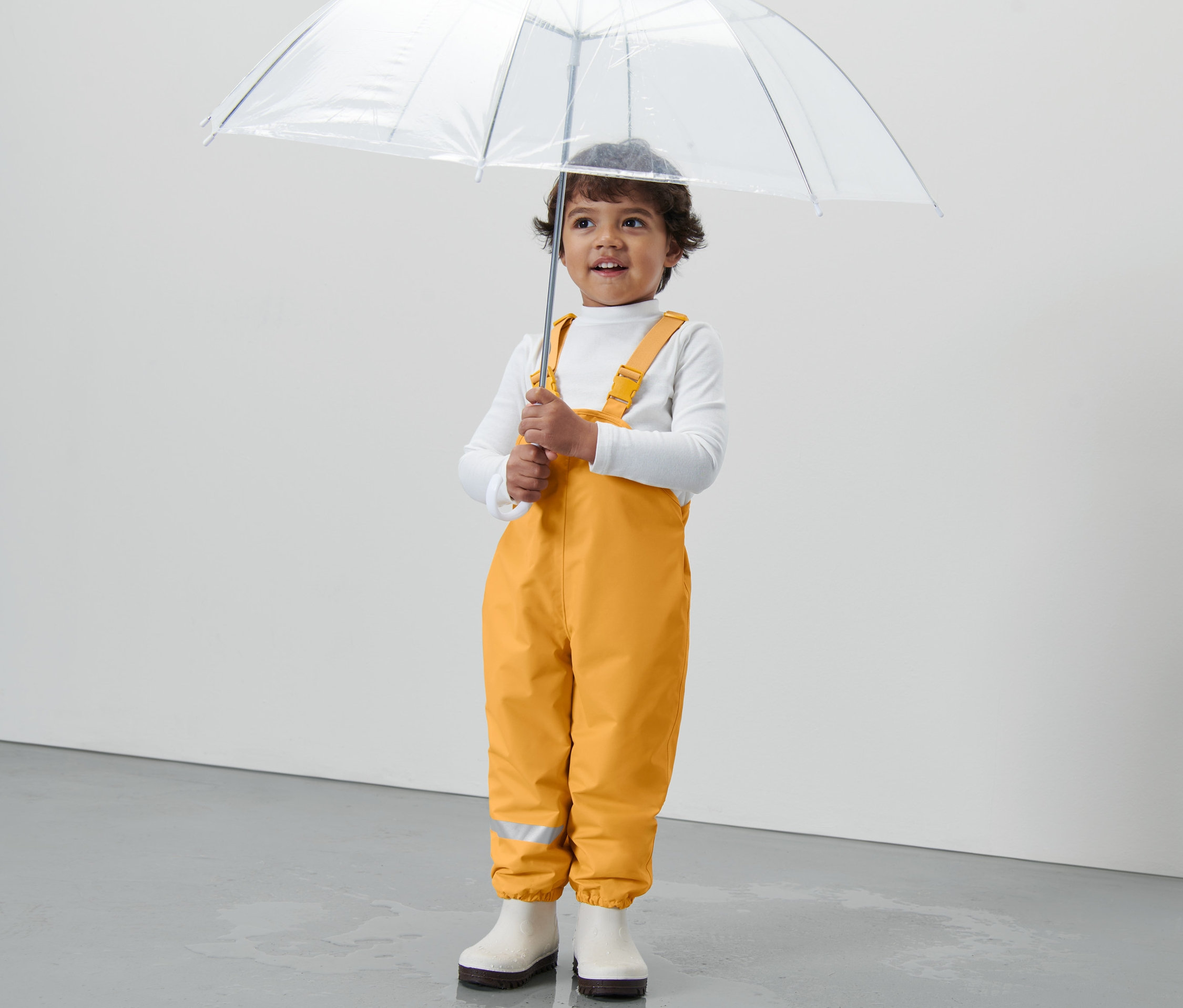Egy gyermek átlátszó esernyőt tart, és kukoricasárga gyerek termo esőnadrágot visel.