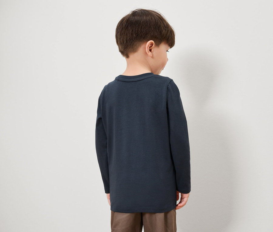 Egy fiú háttal a kamerának, sötétkék hosszú ujjú inget és barna nadrágot visel.