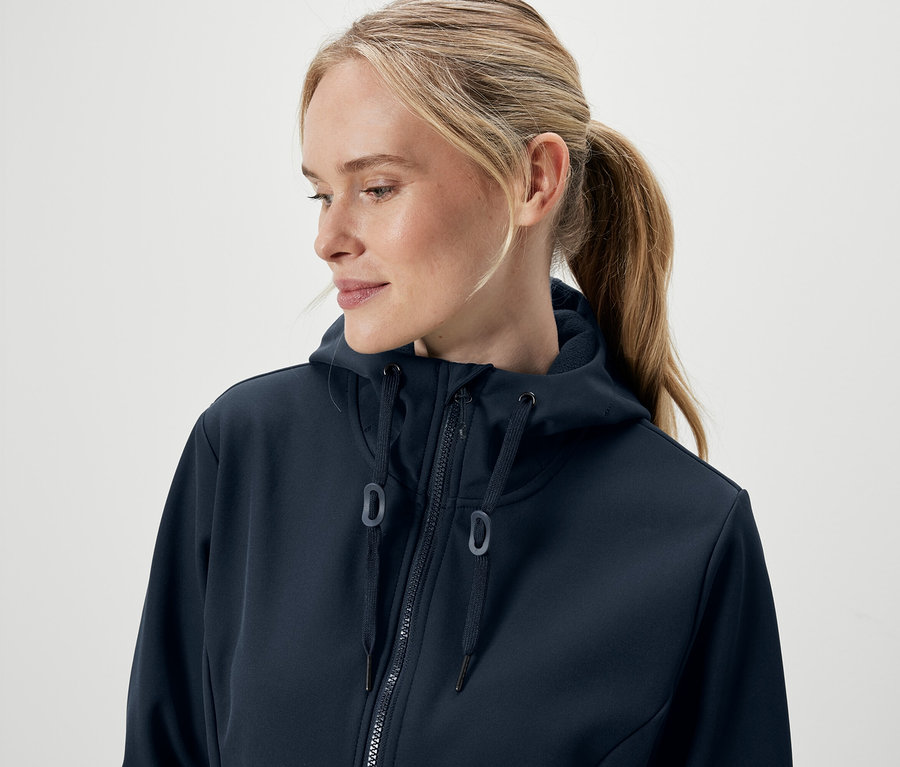 Portré egy szőke hajú, lófarokba kötött nőről, aki sötétkék, cipzáras kapucnis pulóvert visel.