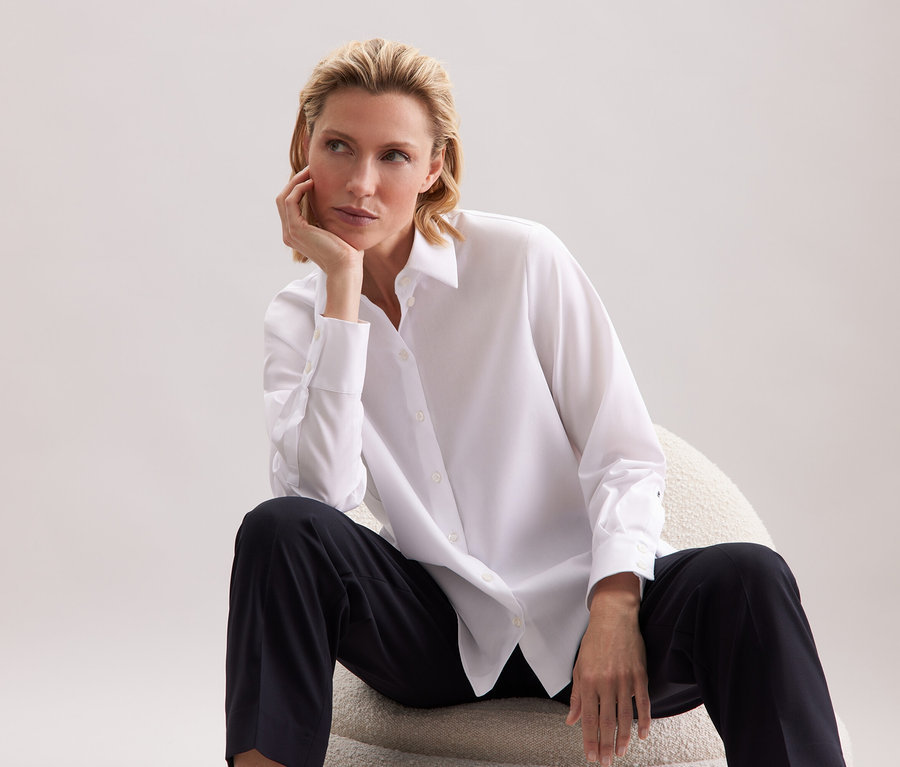 Nő ül egy széken, fehér inget és fekete nadrágot visel.