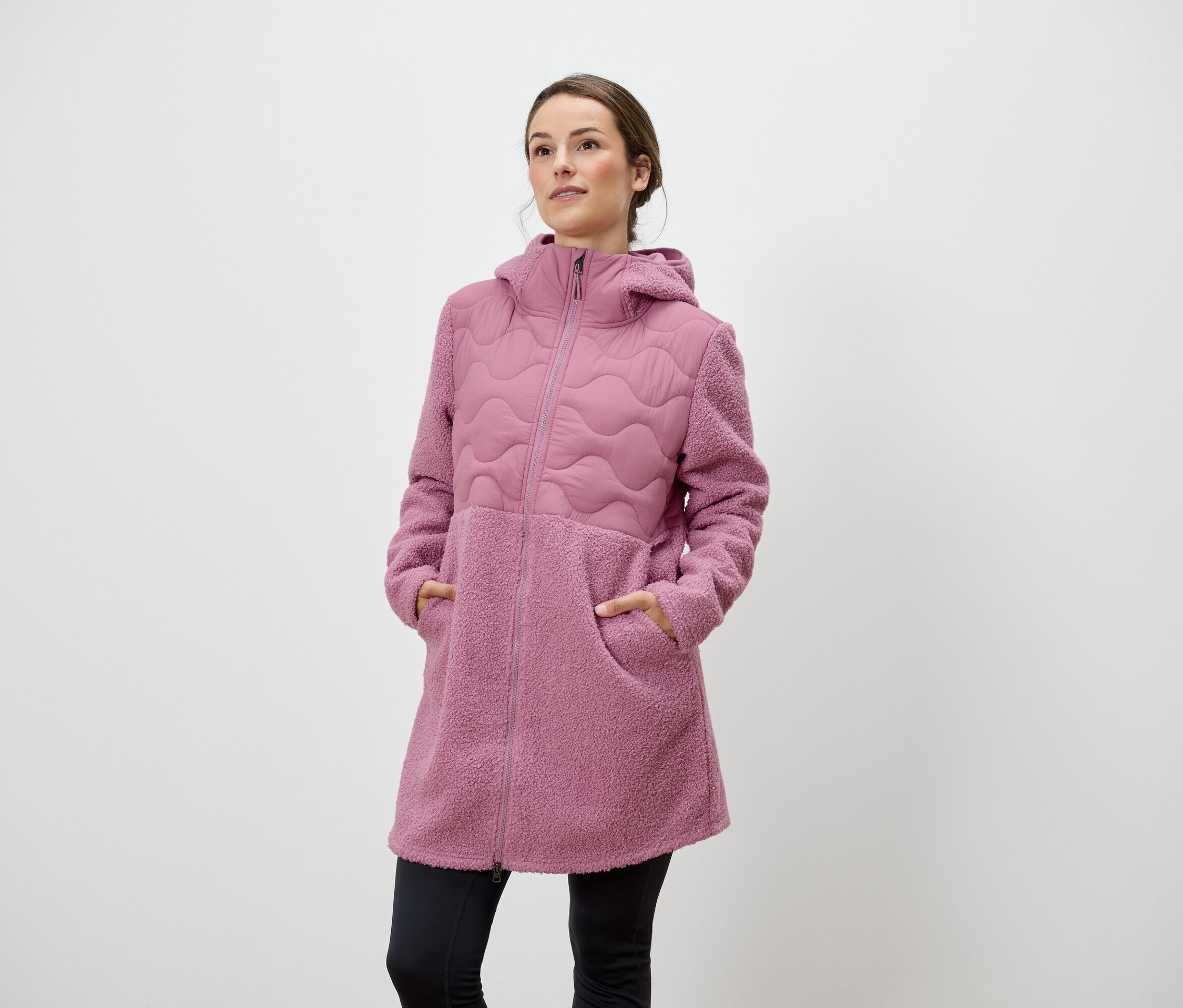 Egy nő áll, kezével egy rózsaszín, cipzáros, kapucnis kabát zsebében.