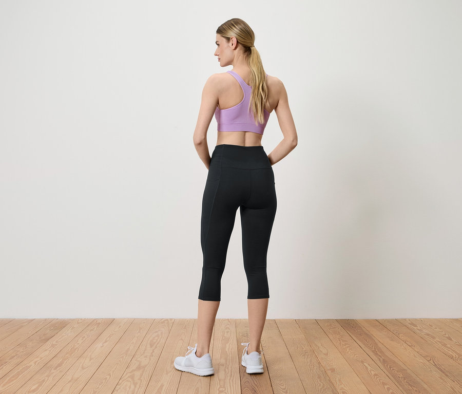 Egy nő háttal áll a kamerának, lila sportmelltartót, fekete leggingset és fehér tornacipőt visel.