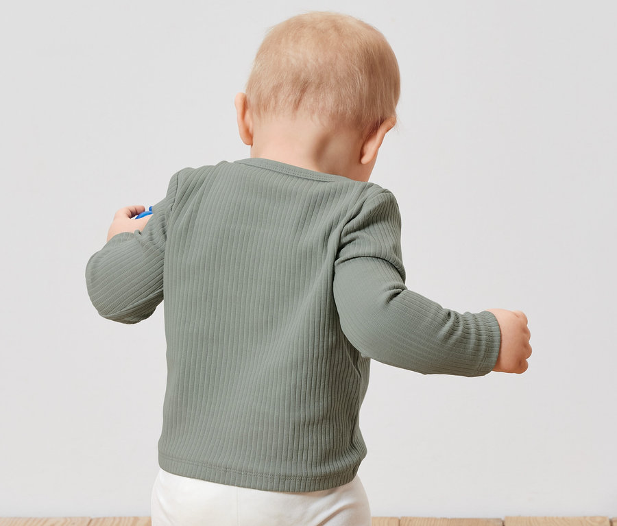 Egy kisgyermek hátulról világoszöld, bordázott, hosszú ujjú felsőt és fehér nadrágot visel.