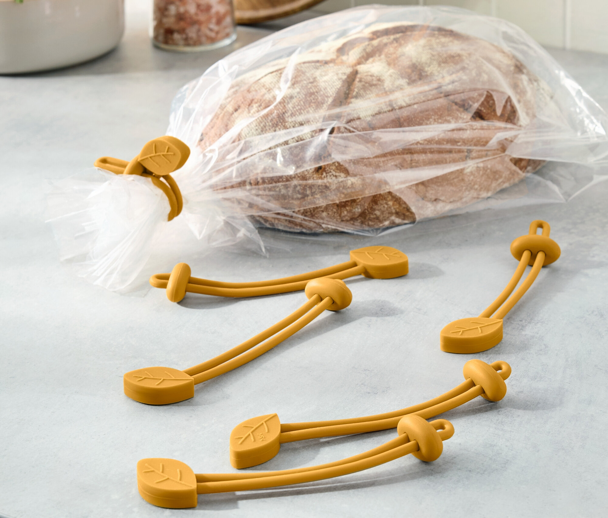 Egy kenyér egy műanyag zacskóban van lezárva egy sárga, levél motívumú gumiszalaggal. Három további gumiszalag van mellette.