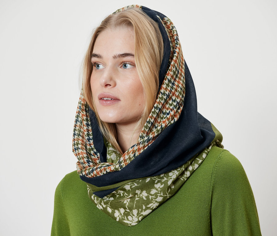 Szőke nő kendőt visel tyúkláb mintával, tengerészkék és virágos réteggel, valamint egy zöld pulóvert.