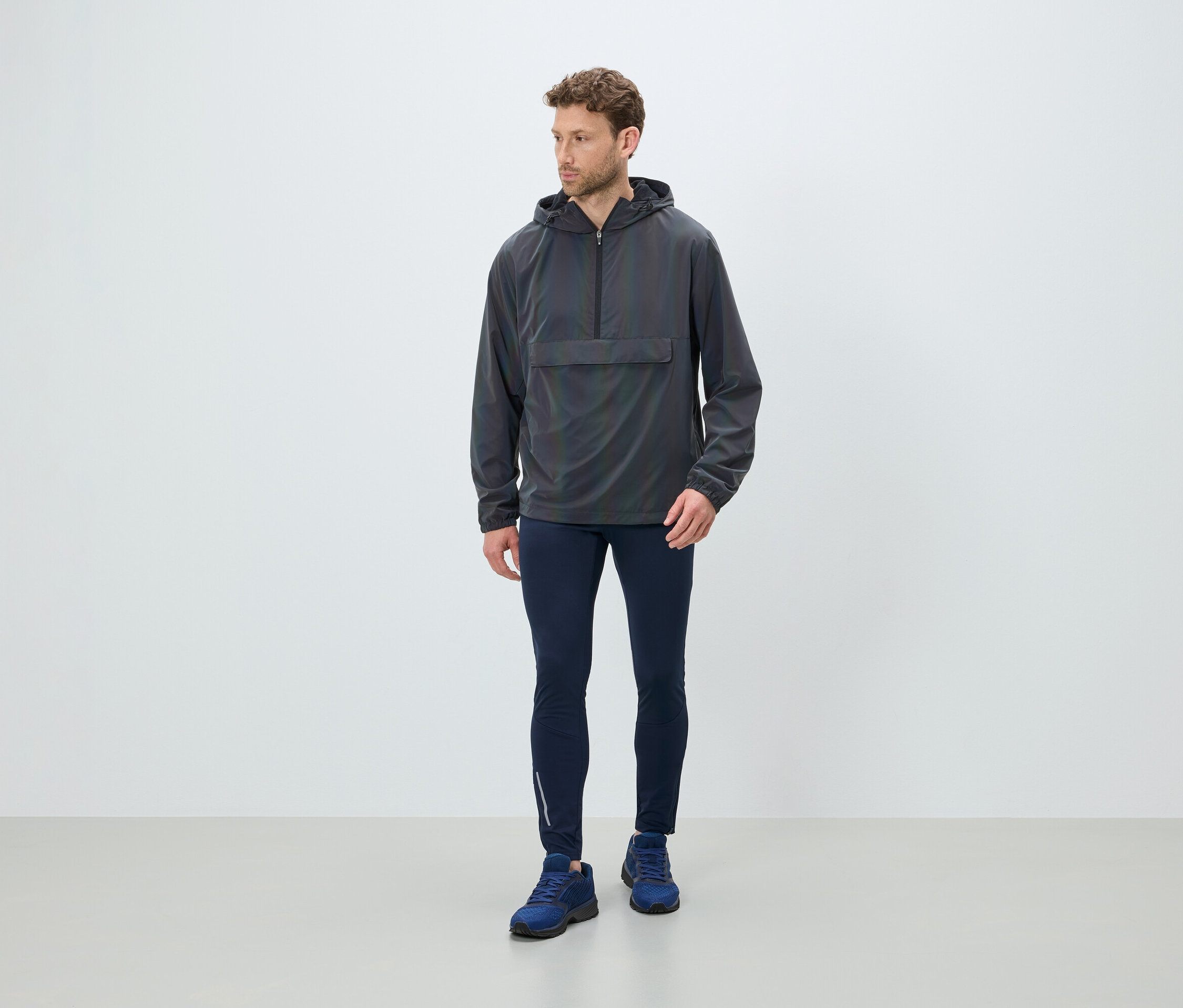 Férfi áll szürke kapucnis széldzsekiben, sötétkék leggingsben és kék cipőben.