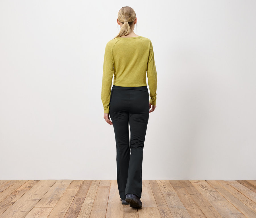 A szőke hajú, lófarokba kötött nő háttal áll a kamerának, olívazöld hosszú ujjú felsőt és fekete nadrágot visel.