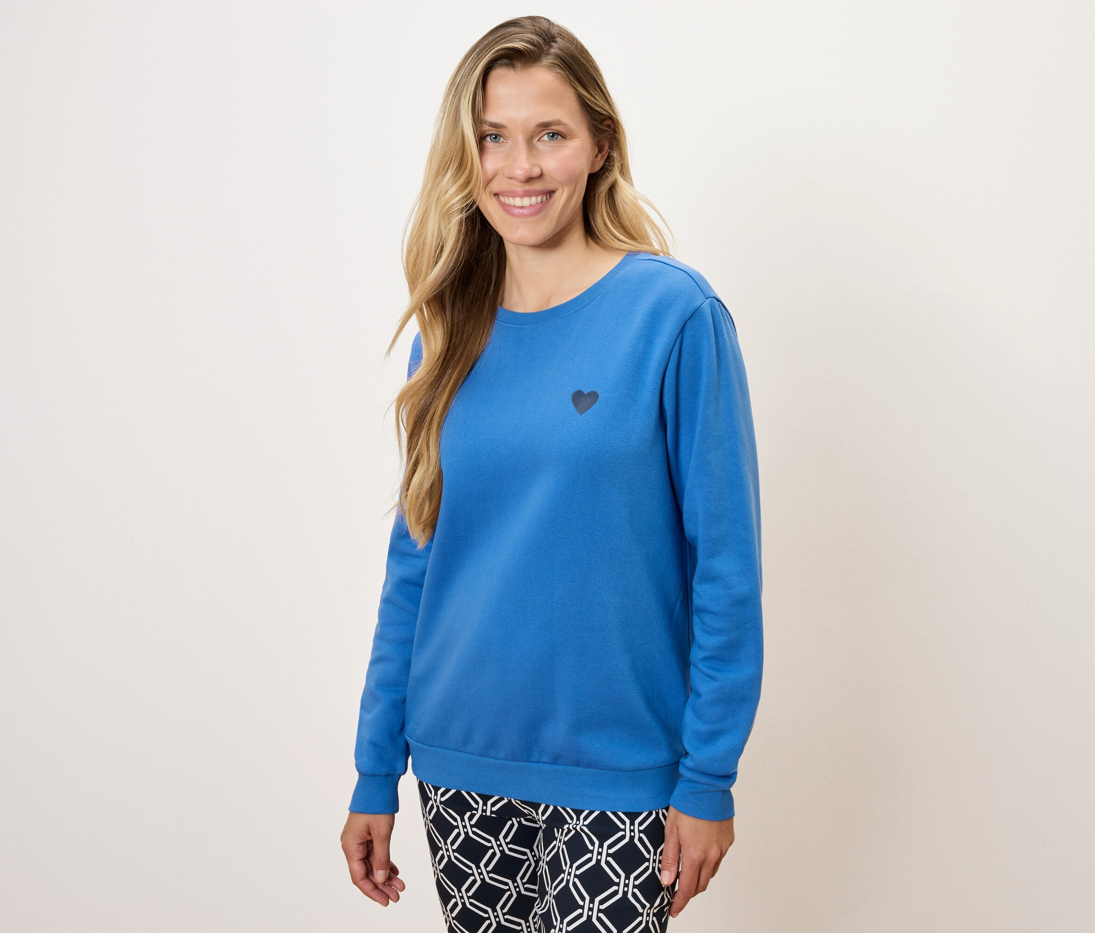 Nő kék, hosszú ujjú pulóvert visel, kis fekete szívvel és mintás nadrágot.