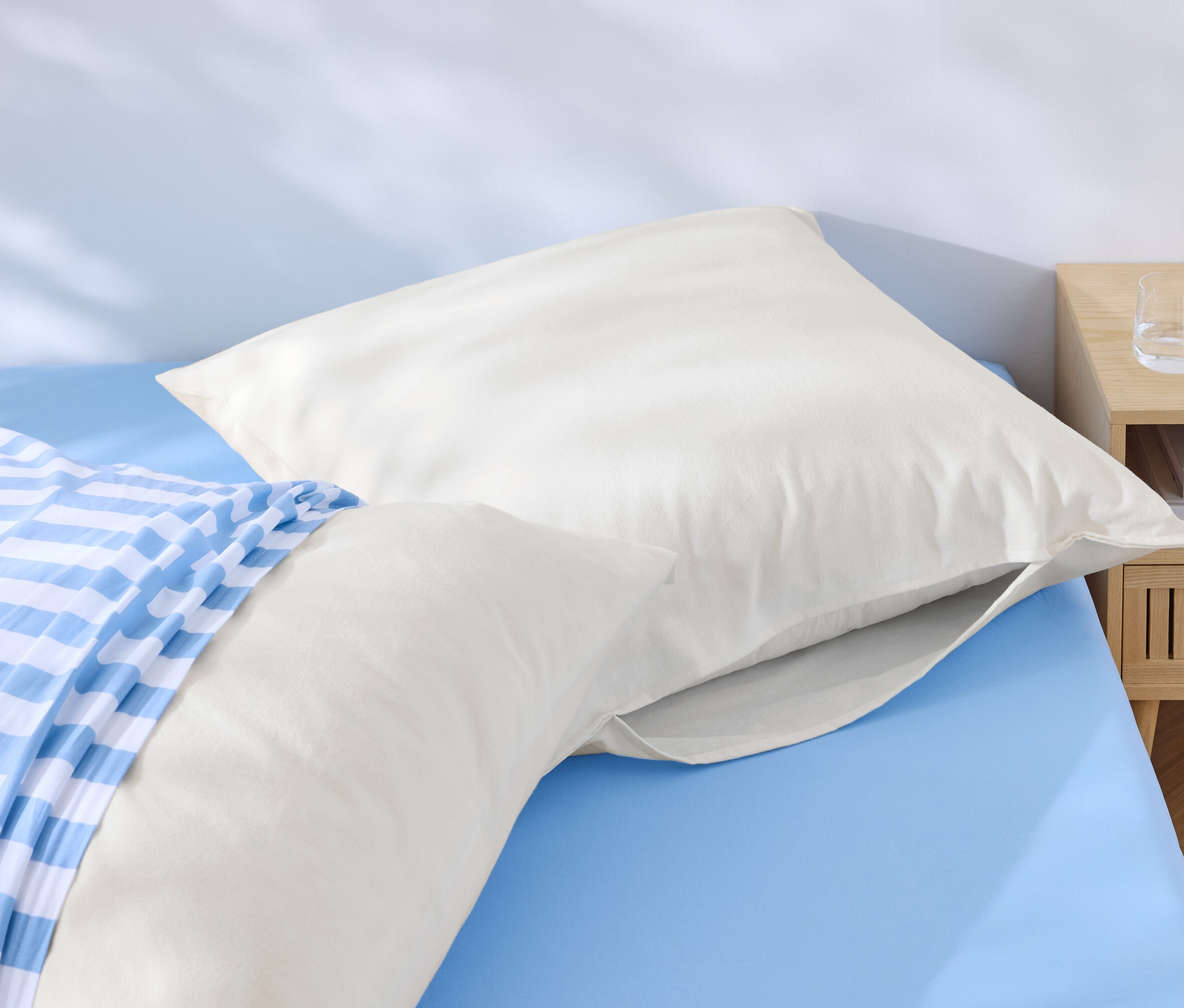 Két fehér párna fekszik egy kék ágyon, az egyik kék-fehér csíkos huzattal. Az ágy mellett egy éjjeliszekrény áll vízzel és egy telefonnal.