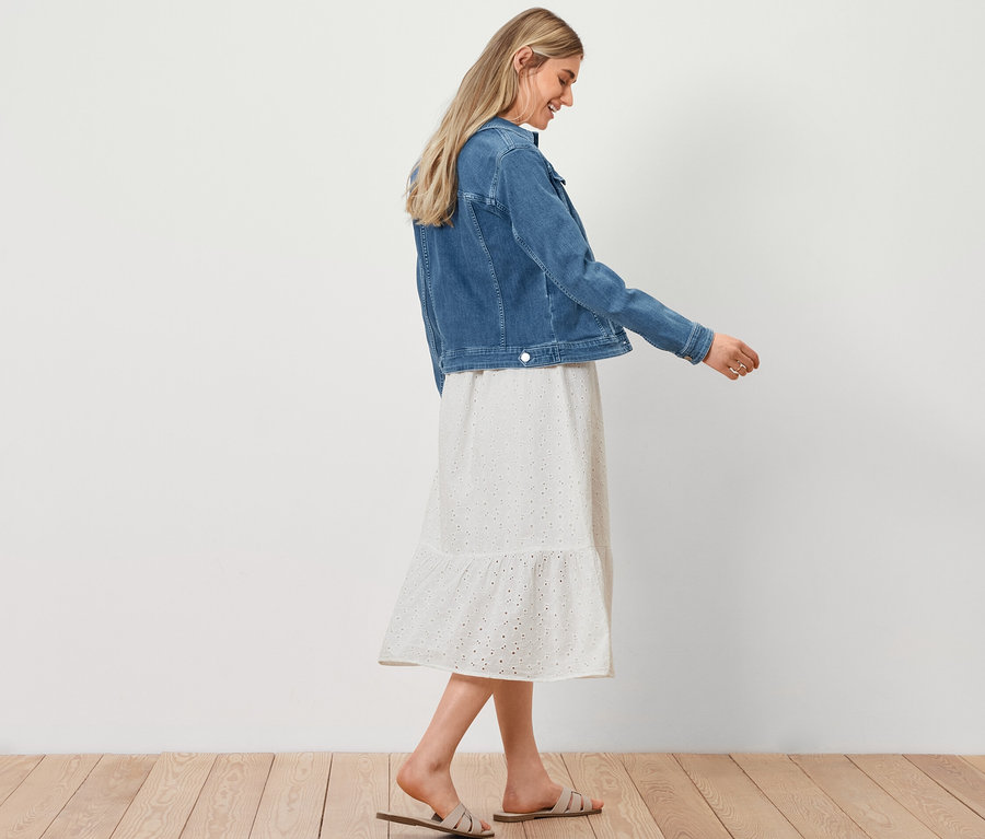 Nő sétál farmerdzsekiben, lyukhímzéses blúzruhában és háncs hatású shopper táskával.