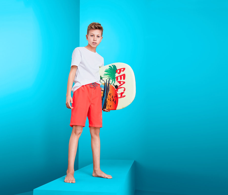 Fiú áll egy emelvényen, egy stranddeszkát tartva, és fiú boardshort fürdőnadrágot visel.