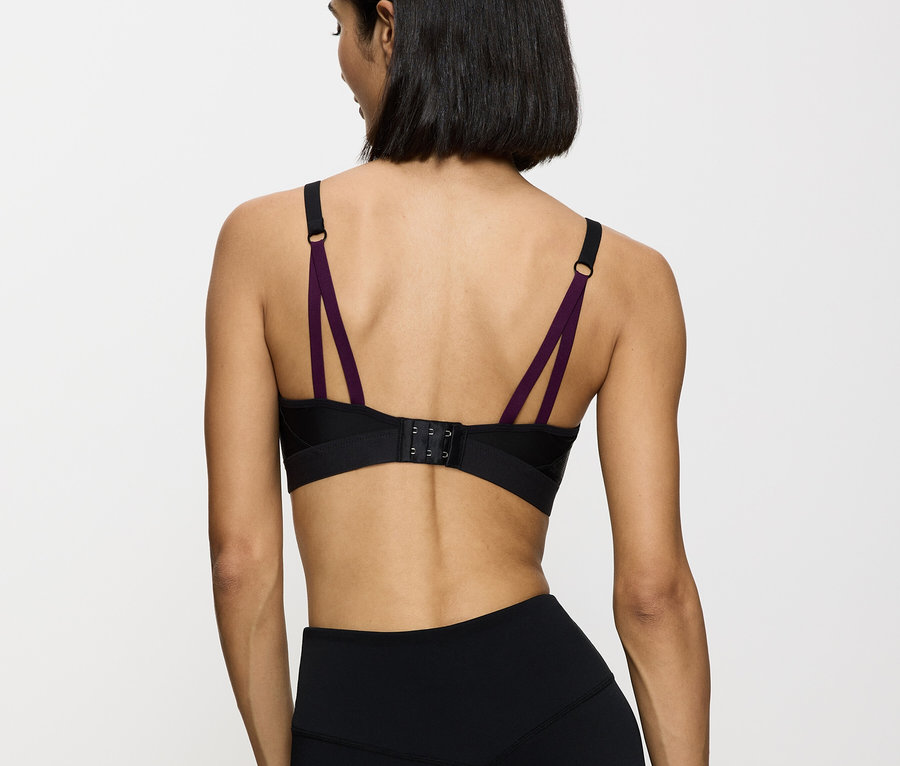 Hátulnézet egy nőről, aki fekete sportmelltartót visel lila pántokkal és fekete leggingset fehér háttér előtt.