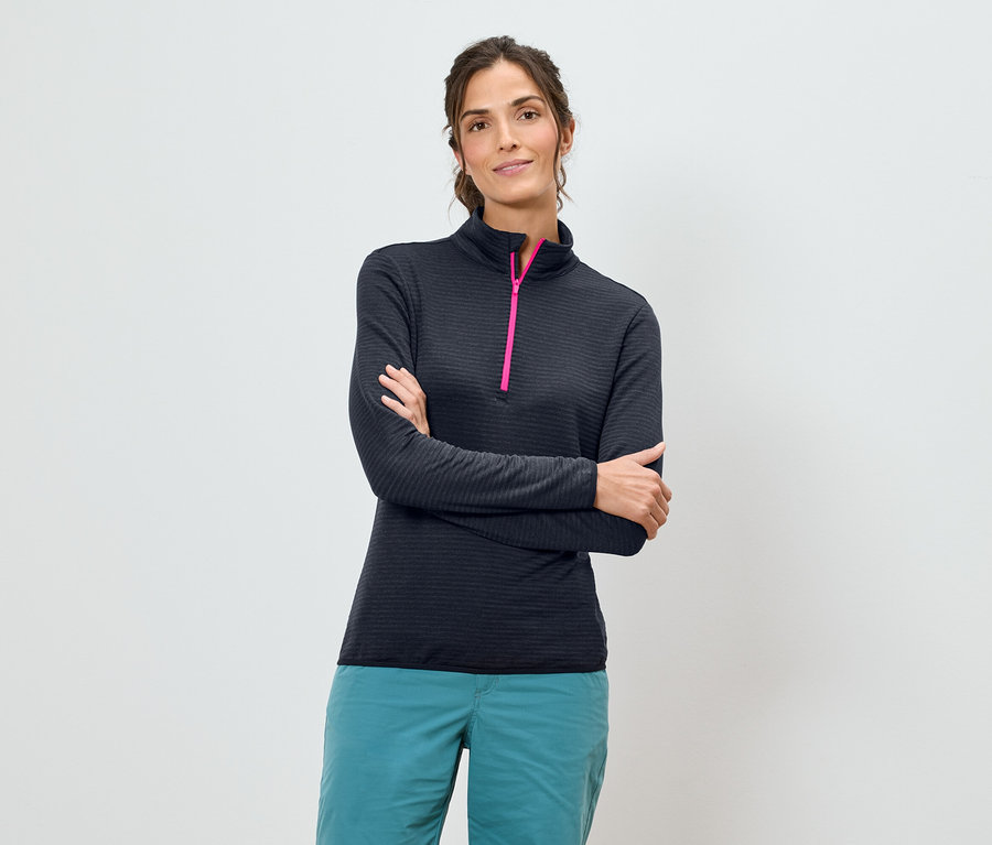 A karjait keresztbe tevő nő sötétkék, hosszú ujjú inget visel rózsaszín cipzárral és türkizkék nadrágot fehér háttér előtt.