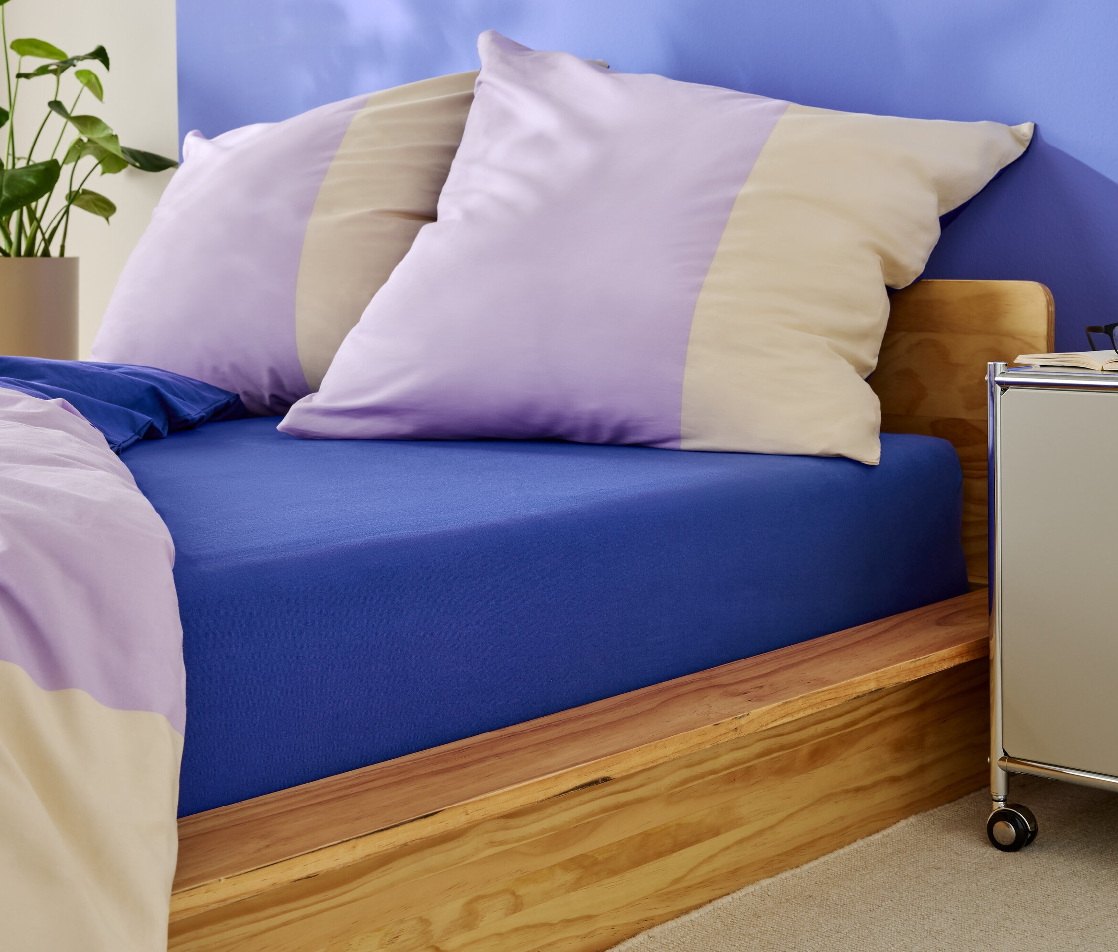 Ágy kék gumis lepedővel és lila és bézs csíkos párnákkal.