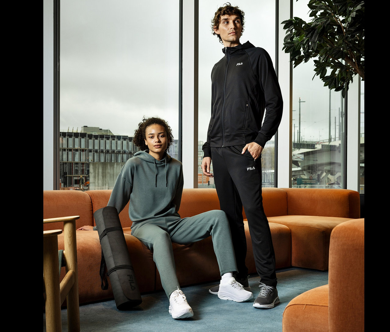 Egy nő ül egy narancssárga kanapén egy fekete jógaszőnyeggel. Mellette egy férfi áll. Mindketten Fila sportruházatot viselnek.