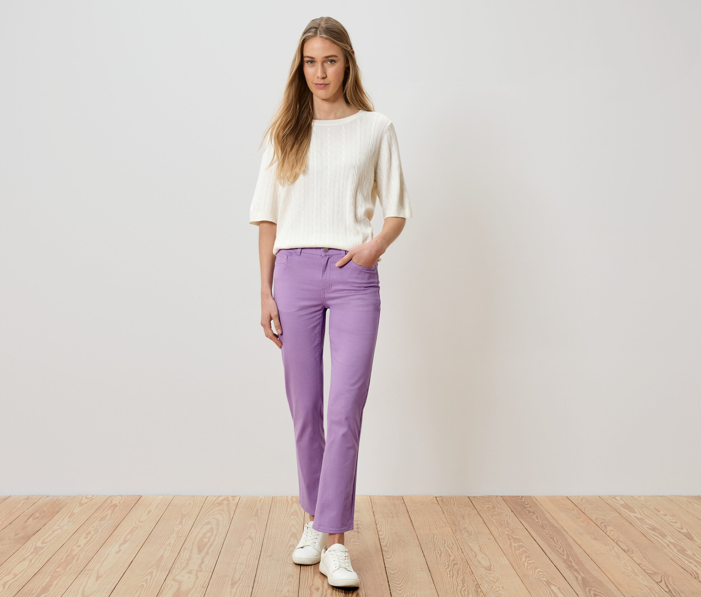 Nő áll, kezével a zsebében, lila nadrágot, fehér pulóvert és fehér cipőt visel.