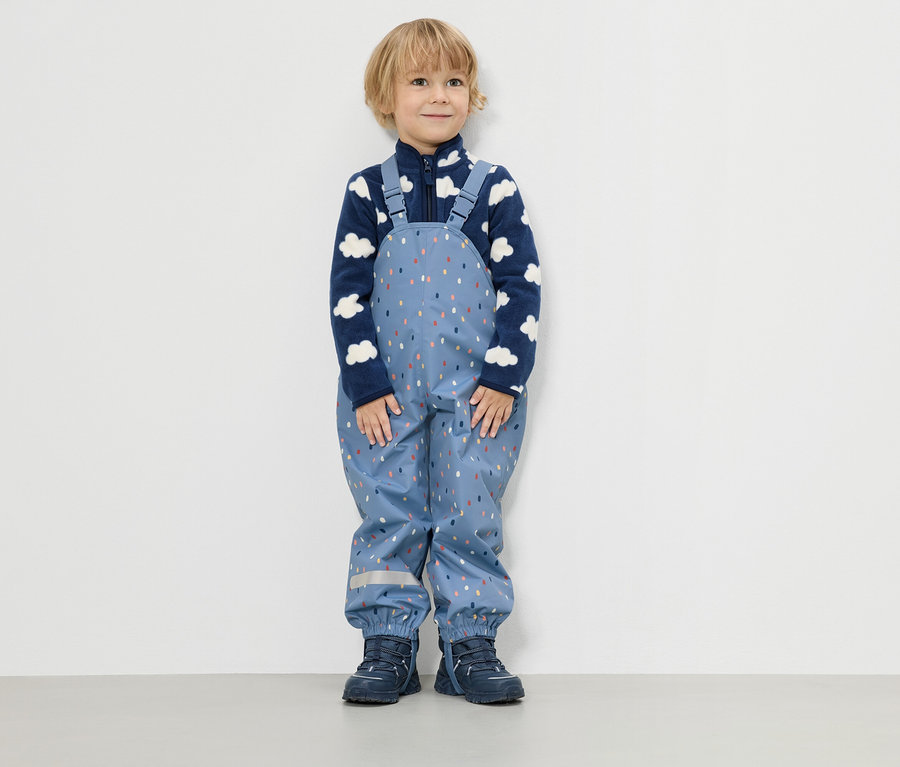 Egy kisfiú áll egy fehér fal előtt, kék, felhőmintás fleece pulóvert és kék, színes cseppmintás esőnadrágot visel.