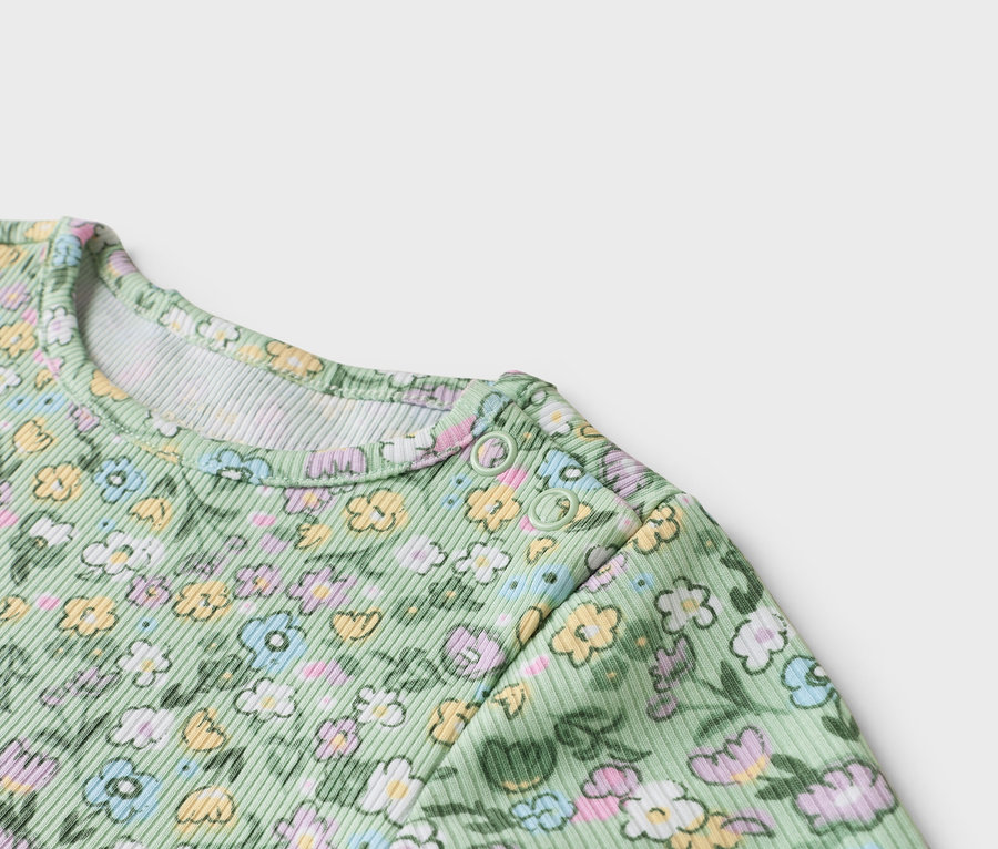 Közeli kép egy virágmintás, világoszöld baba bodyról, melynek a vállán patentok vannak.