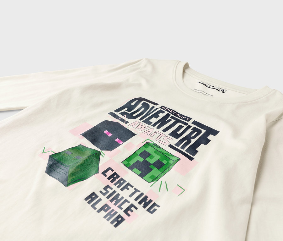 Egy fehér, hosszú ujjú pólón egy Minecraft dizájn látható, a "Adventure Awaits" és a "Crafting Since Alpha" feliratokkal.