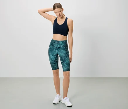 Sportruházatot viselő nő pózol fehér háttér előtt. Sötétkék sportmelltartót és mintás rövid leggingset visel.
