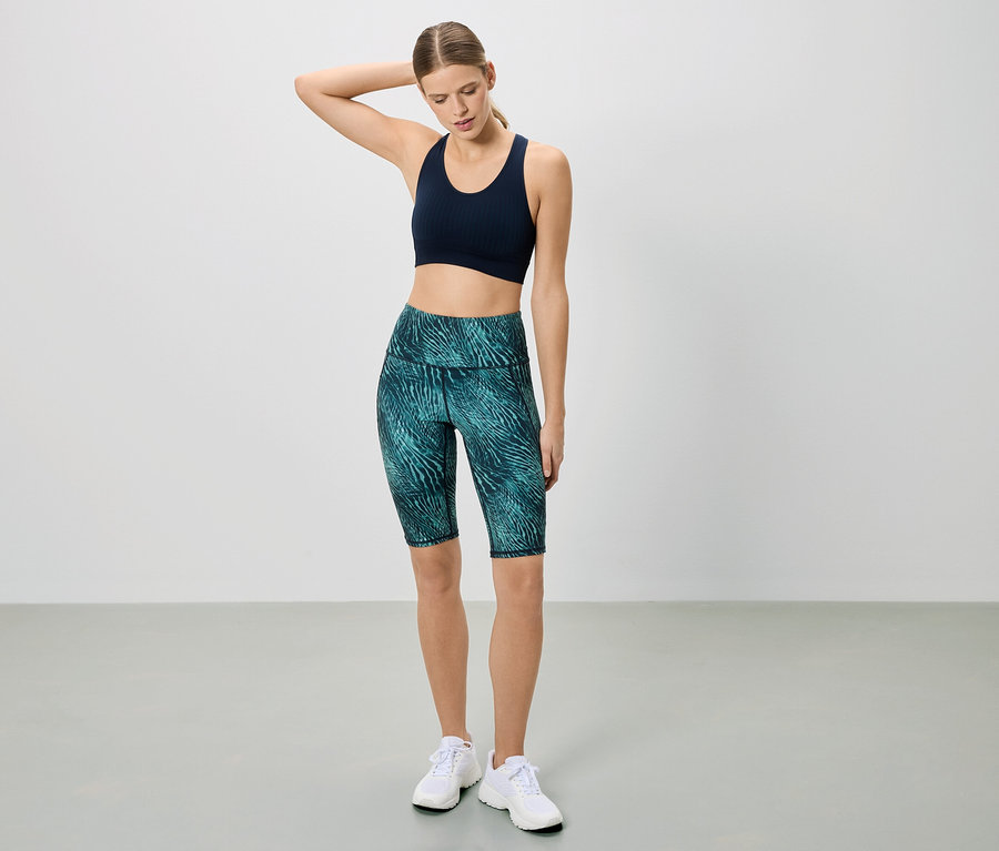 Sportruházatot viselő nő pózol fehér háttér előtt. Sötétkék sportmelltartót és mintás rövid leggingset visel.