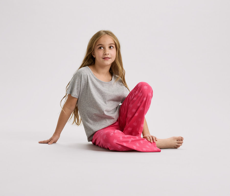 Lány ül a földön, szürke pólót és rózsaszín, pálmafás mintás nadrágot visel.