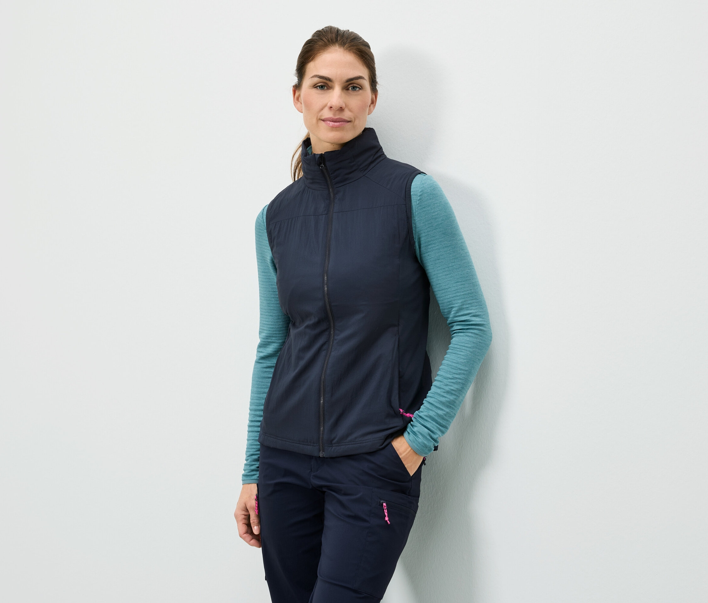 Egy nő áll a fal mellett, kezét a zsebében tartja, sötétkék mellényt, türkizkék hosszú ujjú felsőt és sötétkék nadrágot visel.