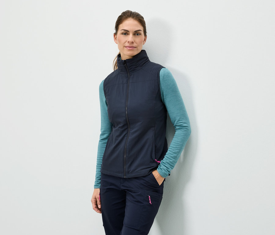 Egy nő áll a fal mellett, kezét a zsebében tartja, sötétkék mellényt, türkizkék hosszú ujjú felsőt és sötétkék nadrágot visel.