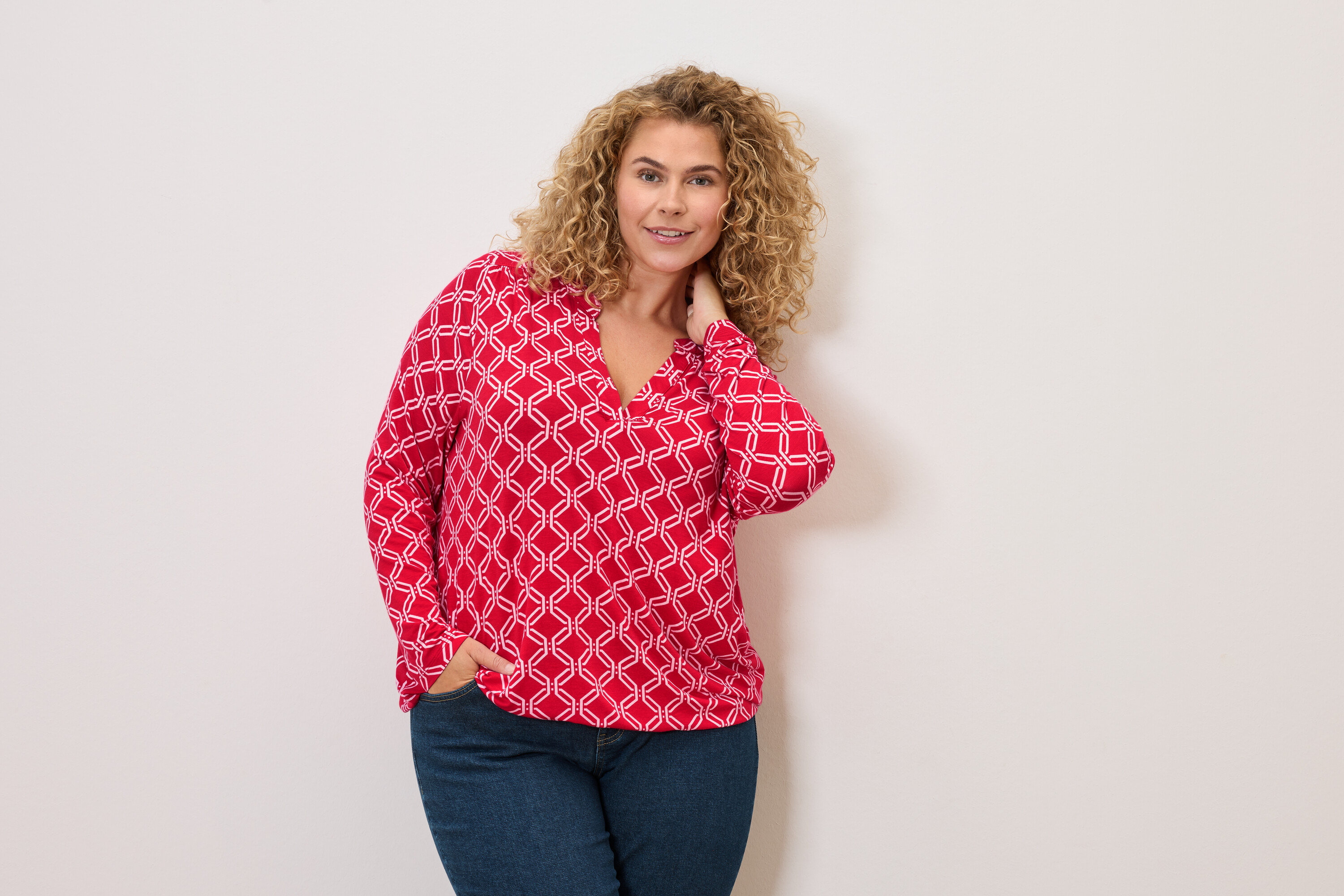 Göndör hajú nő piros-fehér blúzban és farmerben, egyik kezével a zsebében pózol.