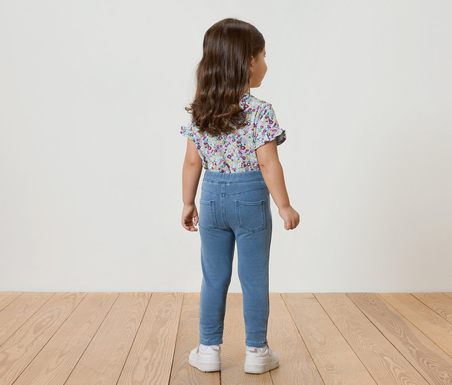 Hátulnézet egy kislányról, göndör hajjal, kék virágos, rövid ujjú felsőben, kék leggingsben és fehér tornacipőben.