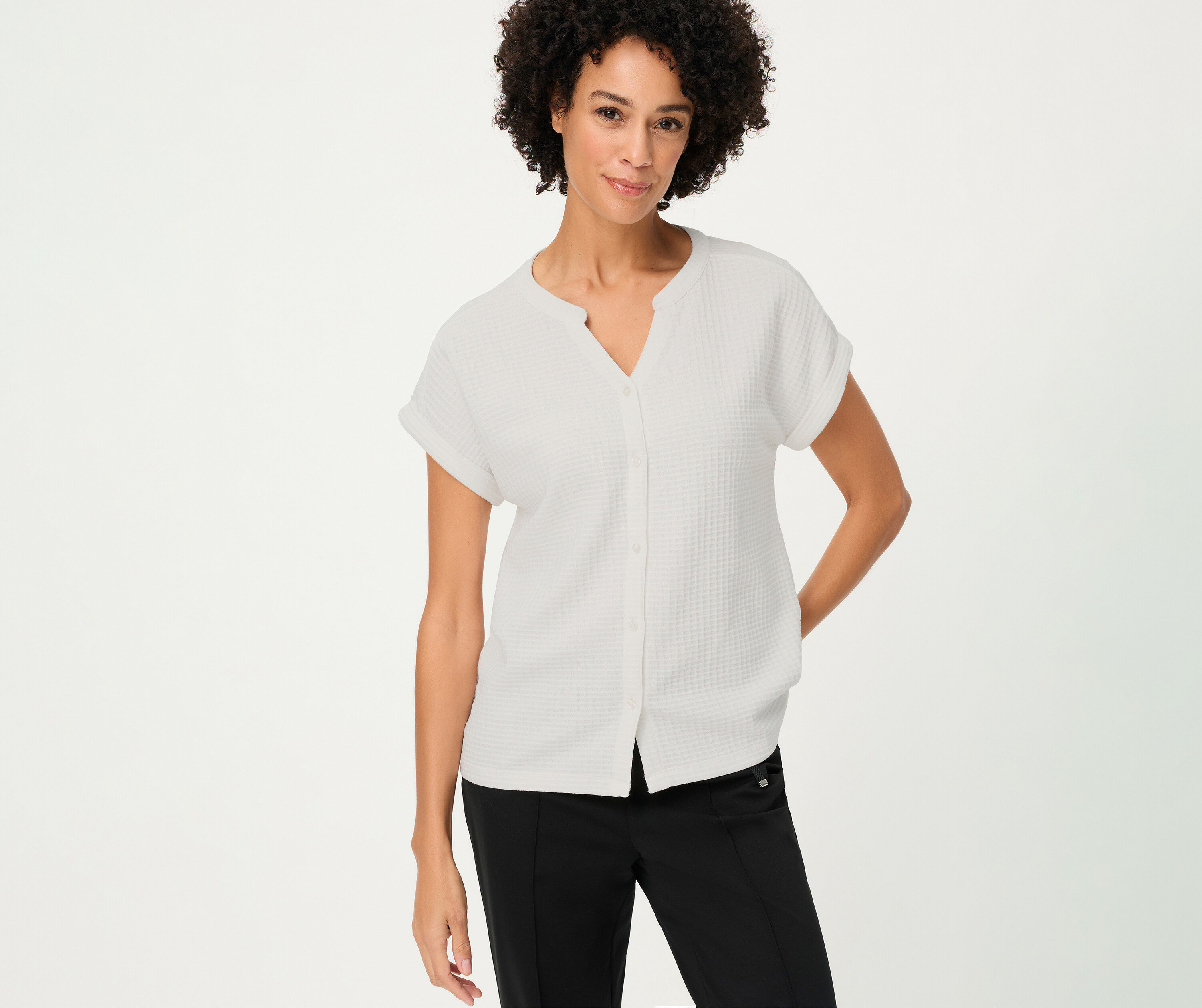 A göndör hajú nő egy fehér, strukturál Olsen pólót visel gombokkal és egy fekete nadrágot.