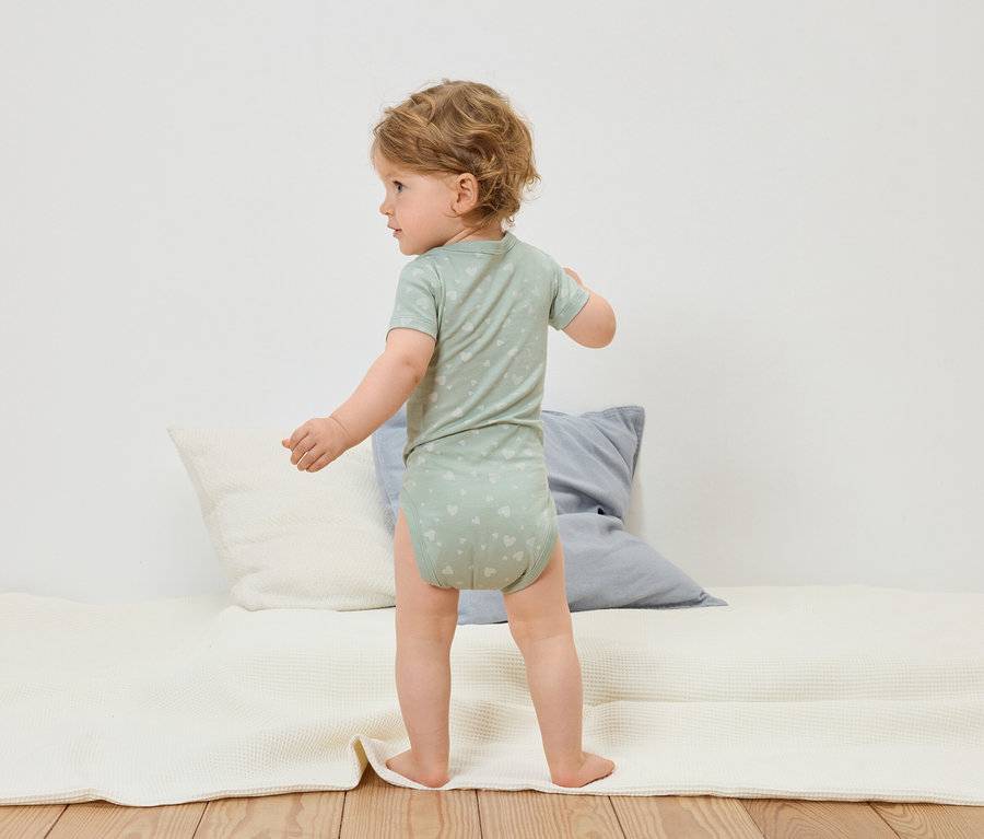 Egy kisgyermek szívmintás, világoszöld bodyban áll egy fehér játszószőnyegen.