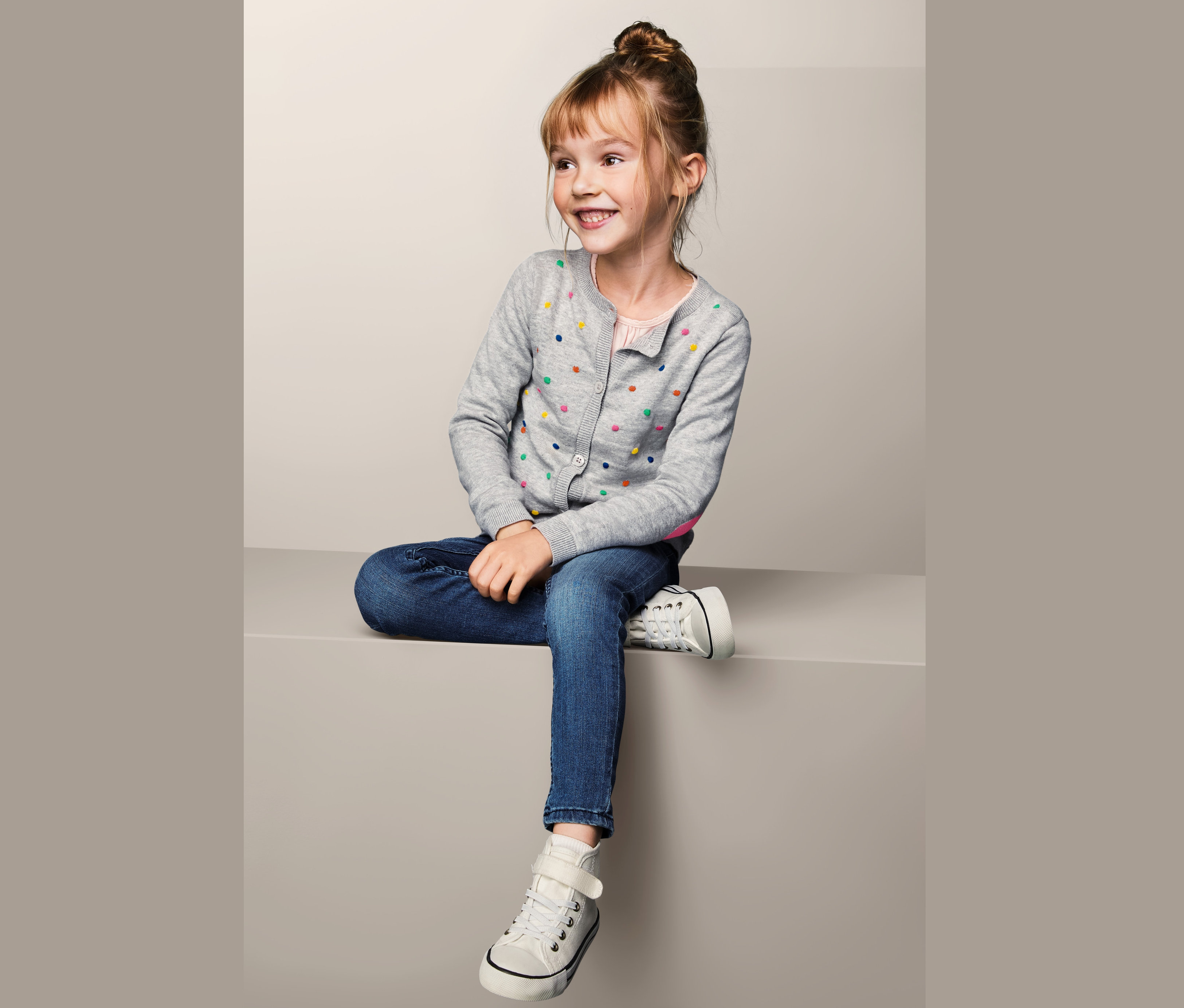 Mosolygó kislány ül kötött kardigánban, farmerben és tornacipőben.