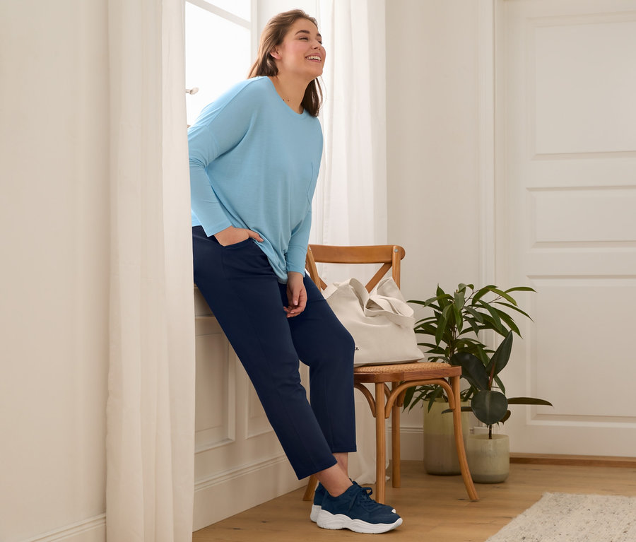 A nő az ablaknak dőlve bőr sneaker cipőt, sötétkék jogging nadrágot, denevérujjú felsőt és vászon bevásárlótáskát visel.