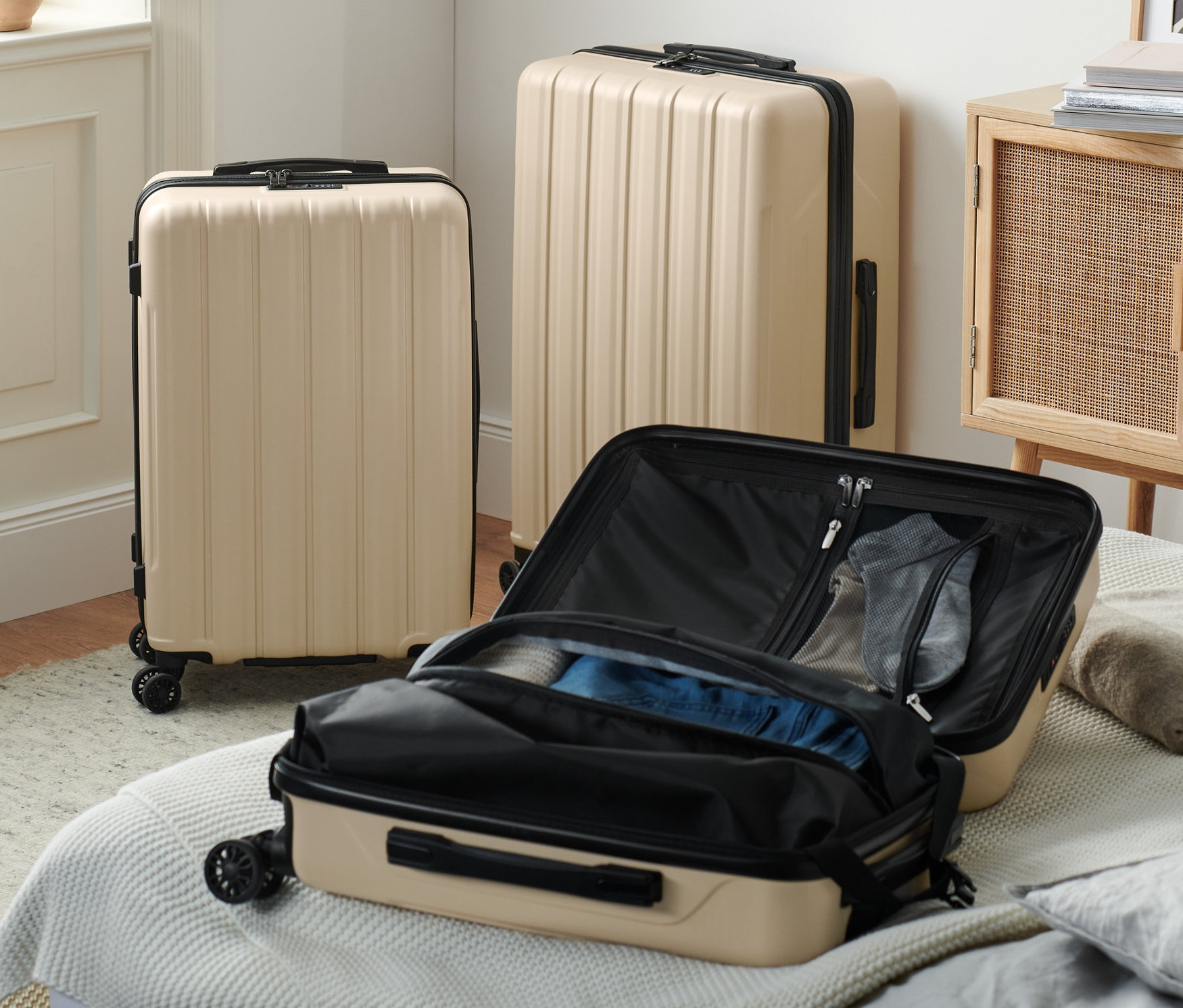 Három bézs keményfedeles bőrönd újrahasznosított anyagból egy belső térben, az egyik nyitva van egy ágyon.