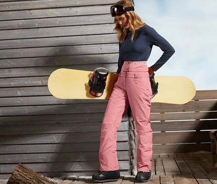 Nő áll egy fal mellett és egy snowboardra támaszkodik. Női sí- és snowboard nadrágot visel, rózsaszín.