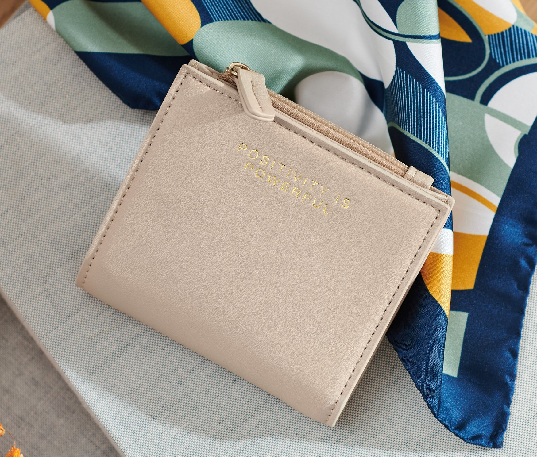 Krémszínű női pénztárca szürke anyagon, virágok és sál mellett.