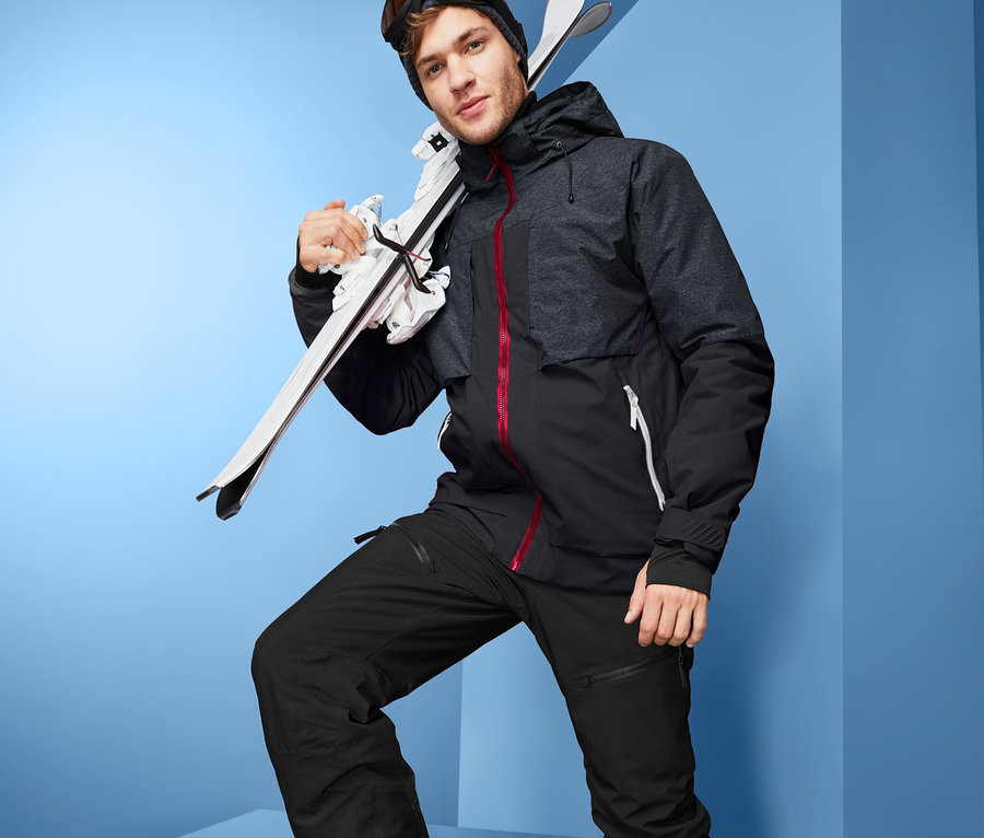 Férfi High-tech síkabátban, sínadrágban, sícipőben és szemüvegben hordja a síléceket a vállán.
