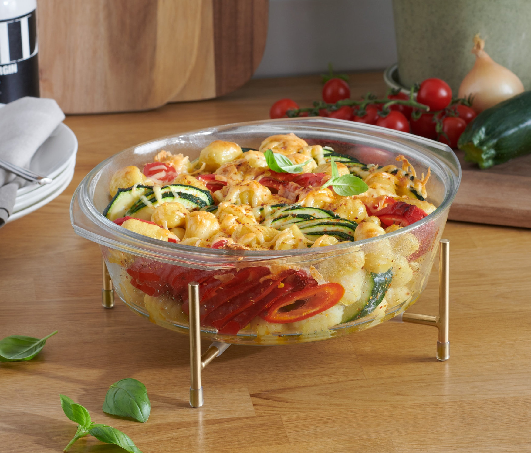 Zöldséges rakott étel üveg sütőformában állvánnyal egy fa asztalon.