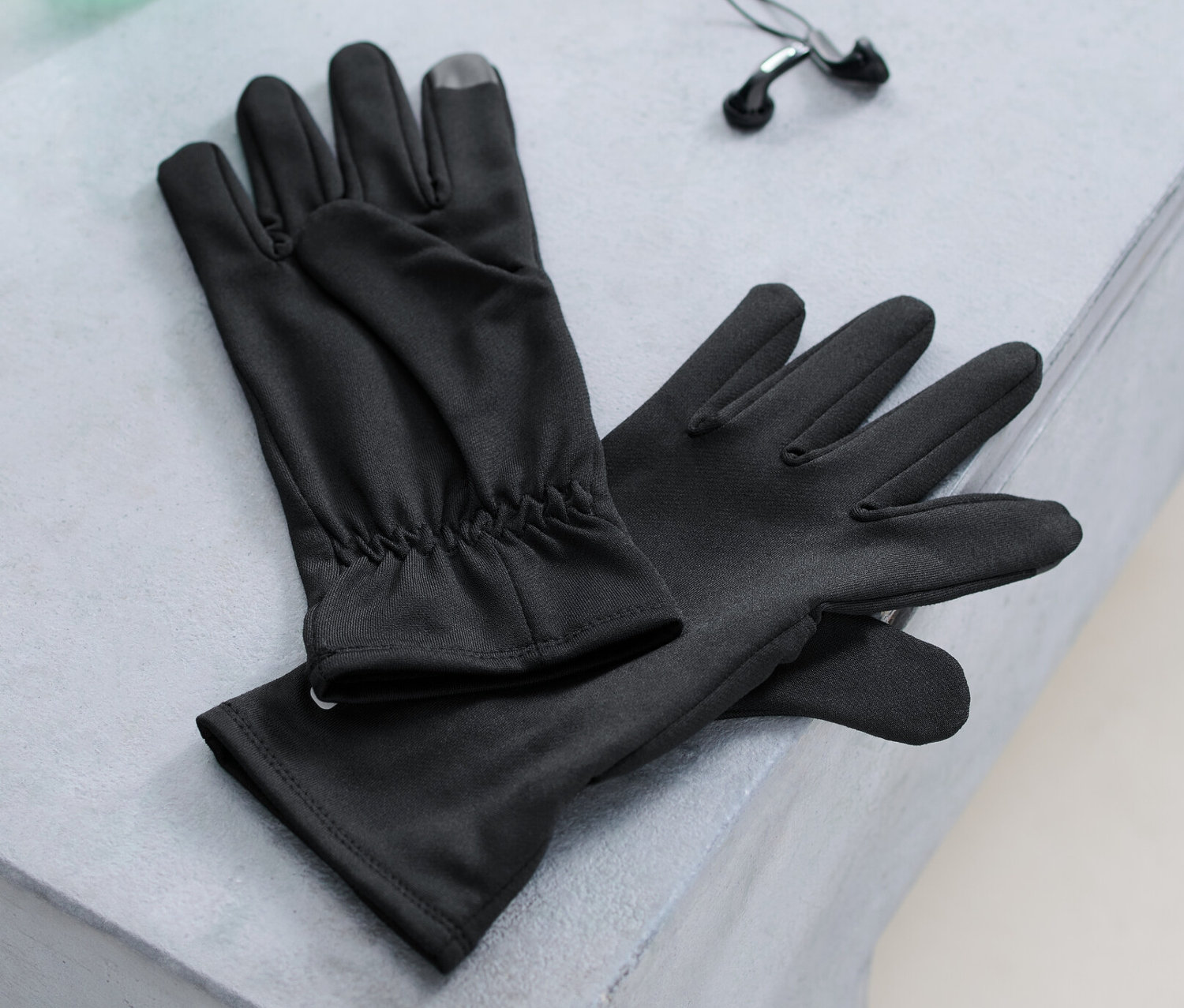 Fekete unisex funkcionális futókesztyű, egy vizes palack és egy fülhallgató fekszik egy padon.