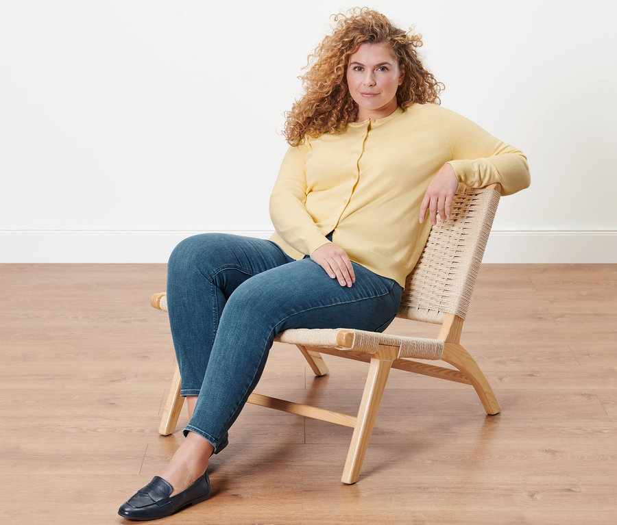 Nő ül a székben, farmer hatású tregging »Fit Nelly« és sárga finomkötésű kardigán van rajta.