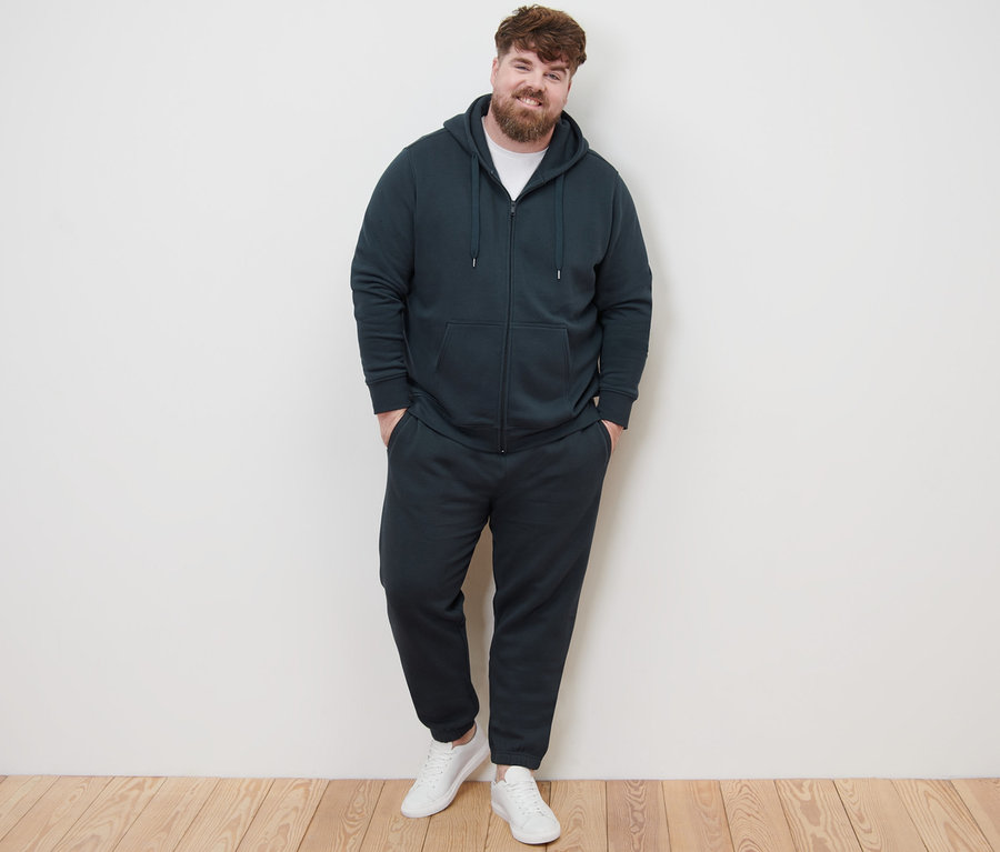 Egy férfi kapucnis szabadidőfelsőben, szabadidőnadrágban és fehér tornacipőben pózol a kamerának.