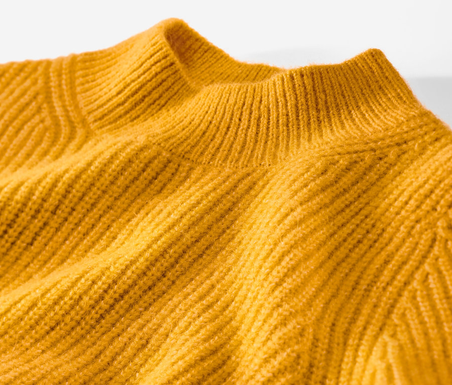 Egy mustársárga, laza kötésű női pulóver részletének képe.