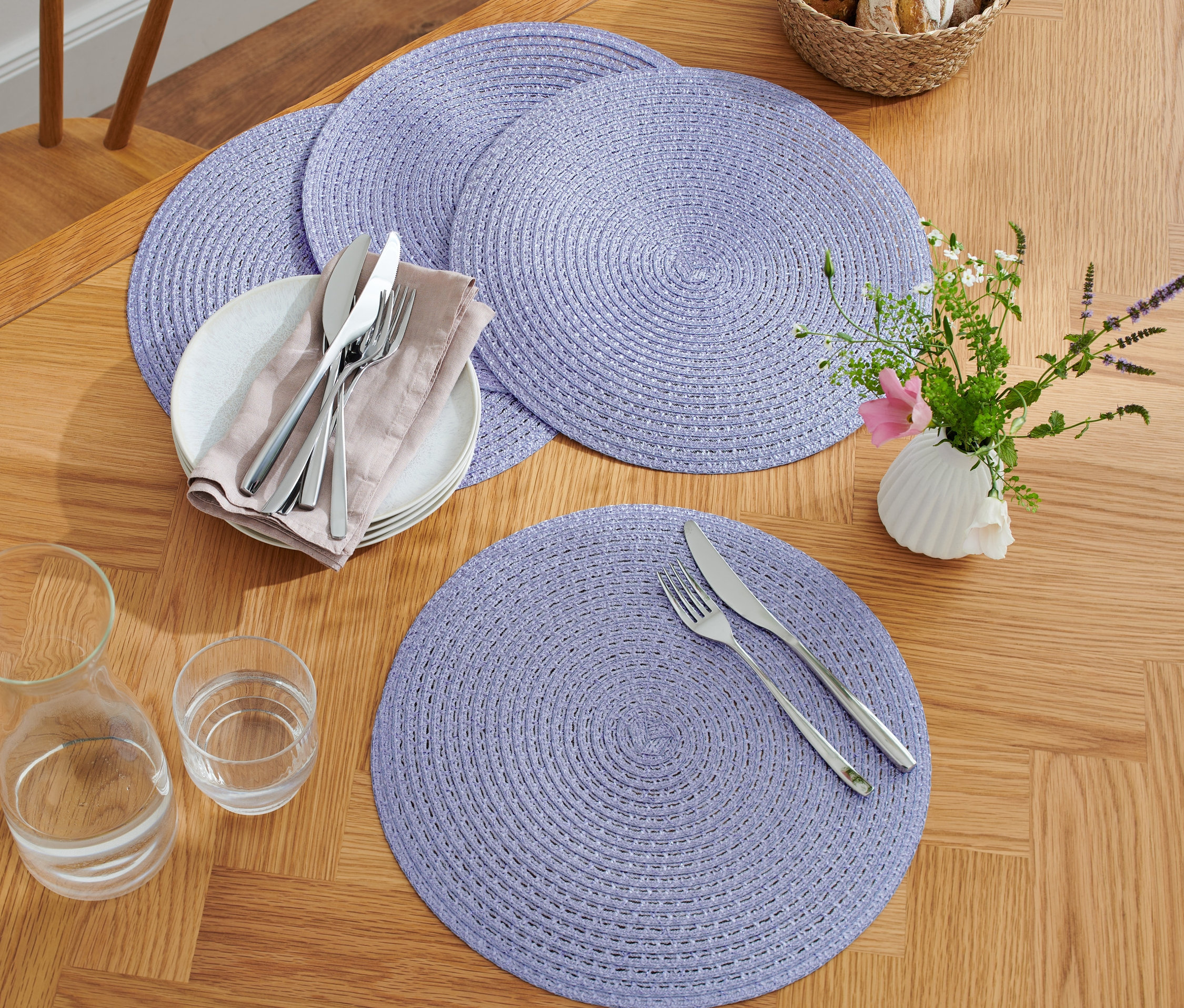 Négy lila alátét egy fa asztalon tányérokkal, evőeszközökkel, poharakkal és egy vázával, virágokkal.