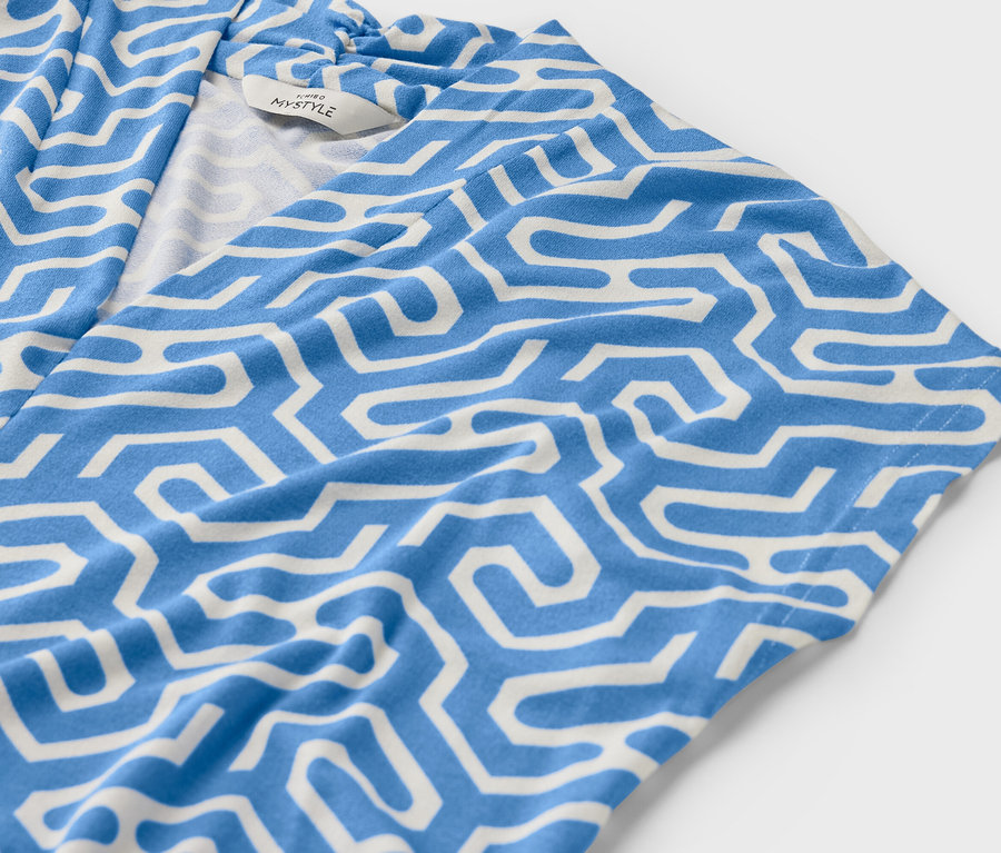 Kék, V-nyakkivágású pólóblúz részlete fehér, geometrikus mintával.