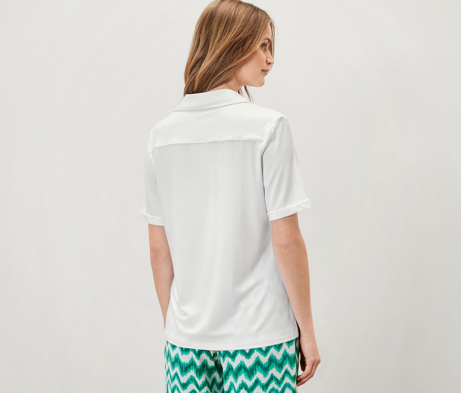 Nő áll háttal a kamerának, fehér női pólóblúzt és zöld mintás nadrágot visel.