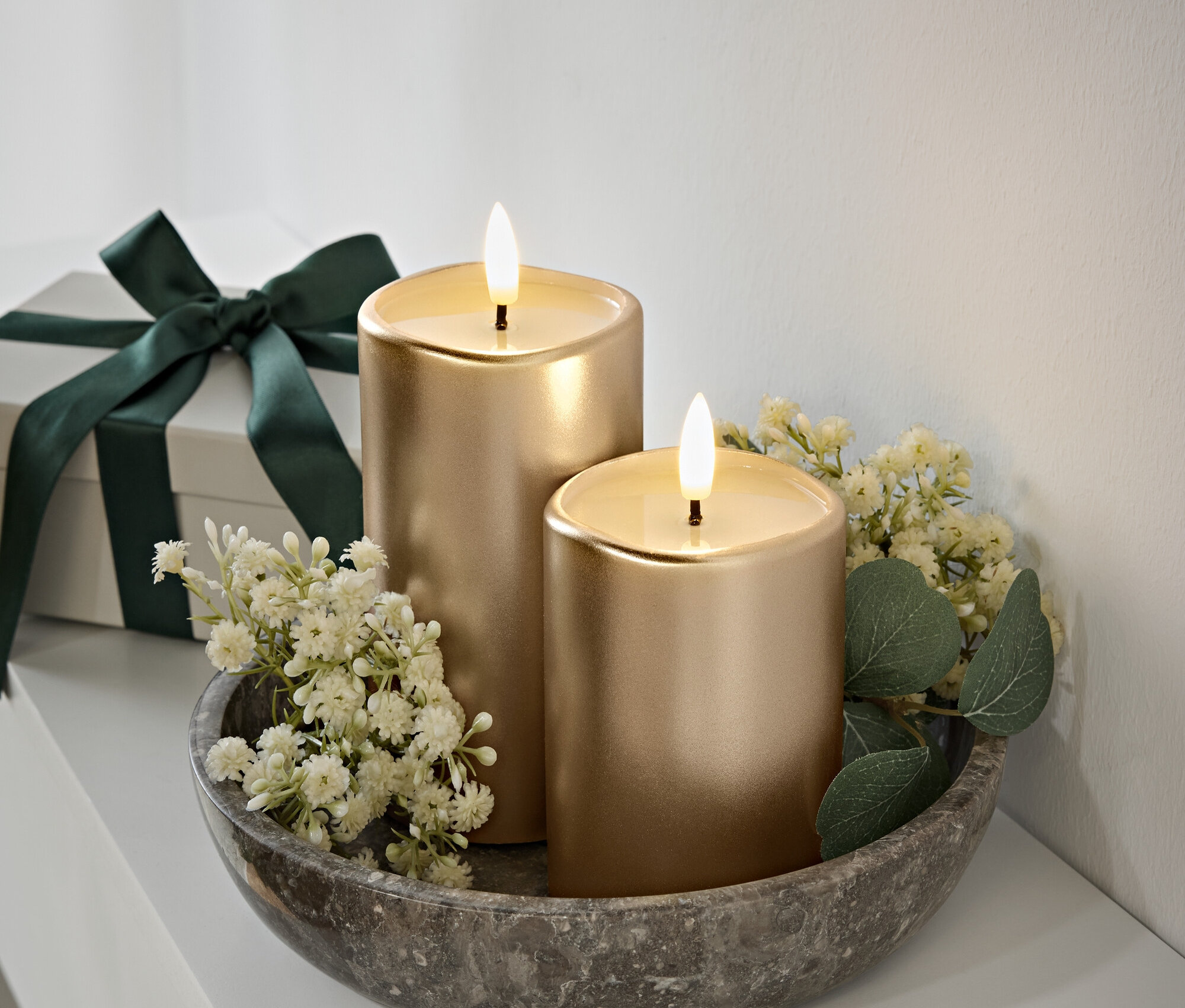 Két arany gyertya lánggal egy szürke kőtálban, fehér virágok és zöld levelek. Ajándékdoboz sötétzöld szalaggal a háttérben.